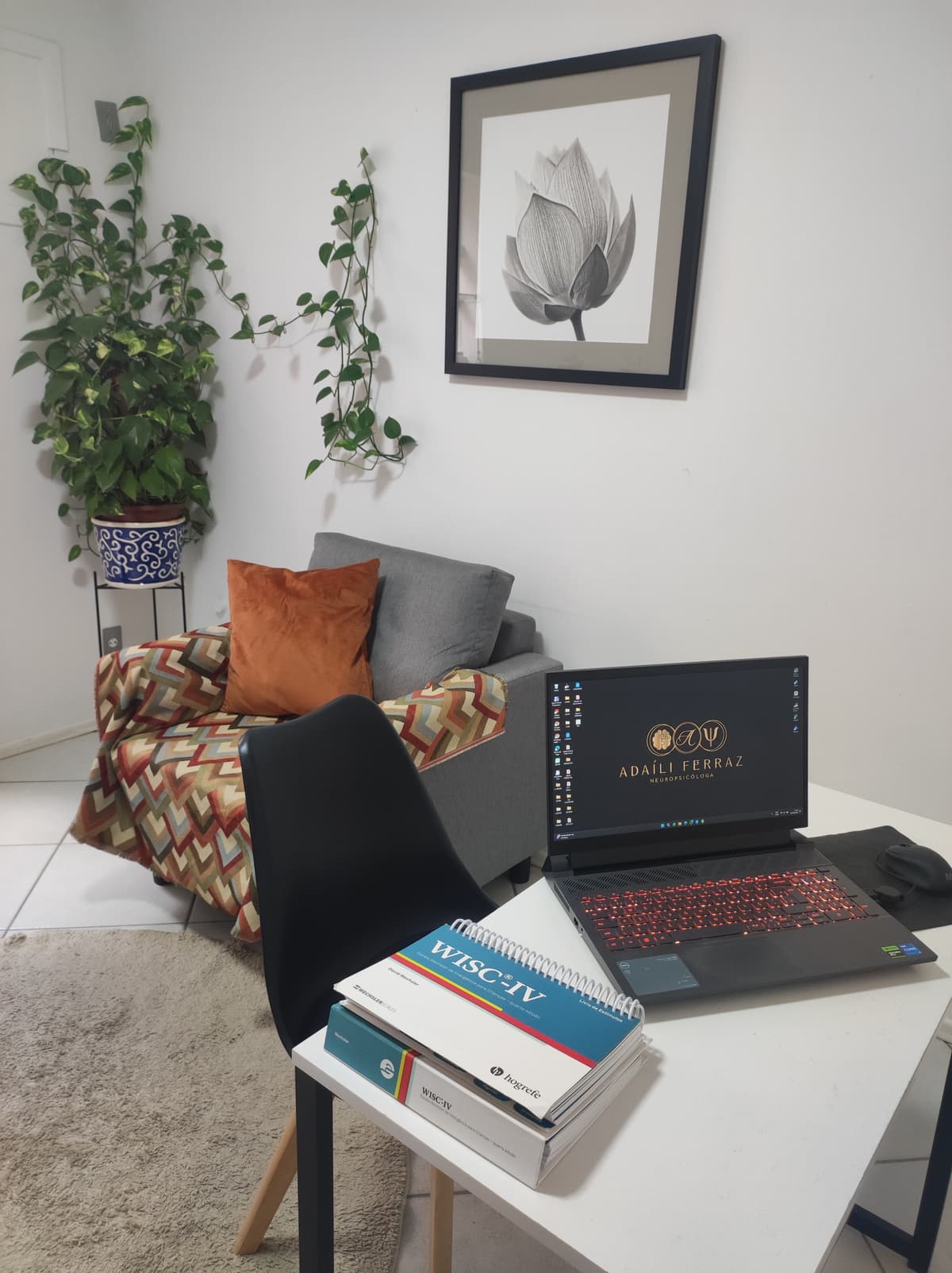 Sala de Psicoterapia com uma poltrona cinza e manta marrom com uma almofada marrom, acima um quadro com uma flor cinza, ao lado direito um vaso com plantas, a direita uma cadeira preta e uma mesa branca com um notebook e testes ao lado